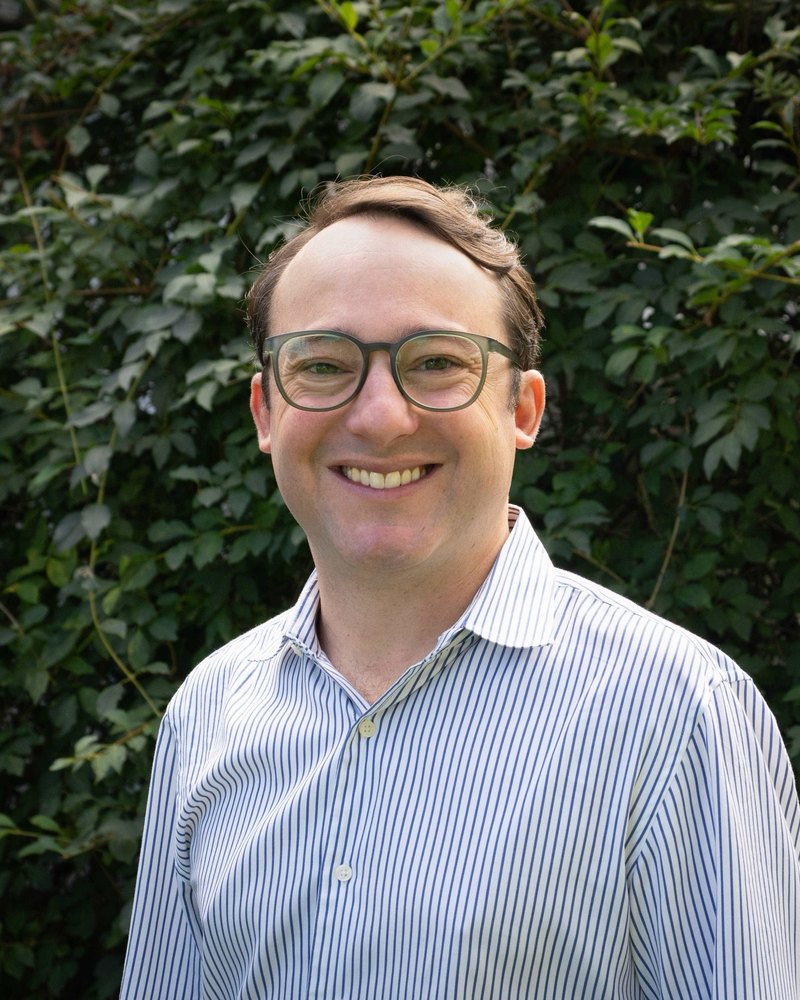 Adam Winski, smiling, in a striped shirt in front of green leaves